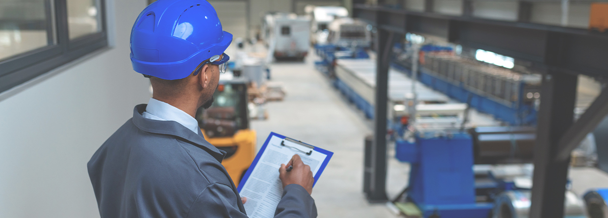 Person in a blue hard hat and safety glasses holds a clipboard while inspecting equipment and machinery inside a large industrial facility.
