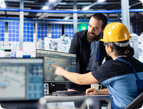 Engineer in a yellow hard hat pointing at a 3D design on a computer screen while discussing with a colleague in a modern industrial workspace.